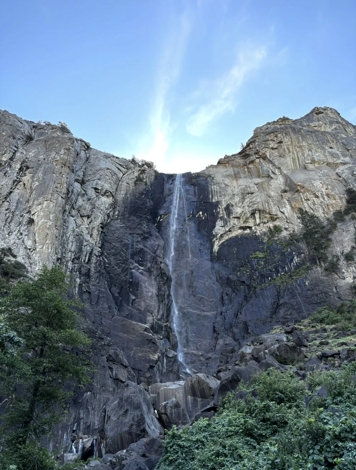 Tall waterfall flowing down a rocky mountain with greenery at the base, clear blue sky above.