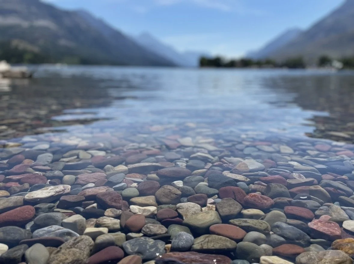 Close-up view of colorful smooth pebbles on a lakebed, with shallow water, mountains, and a blue sky in the background.