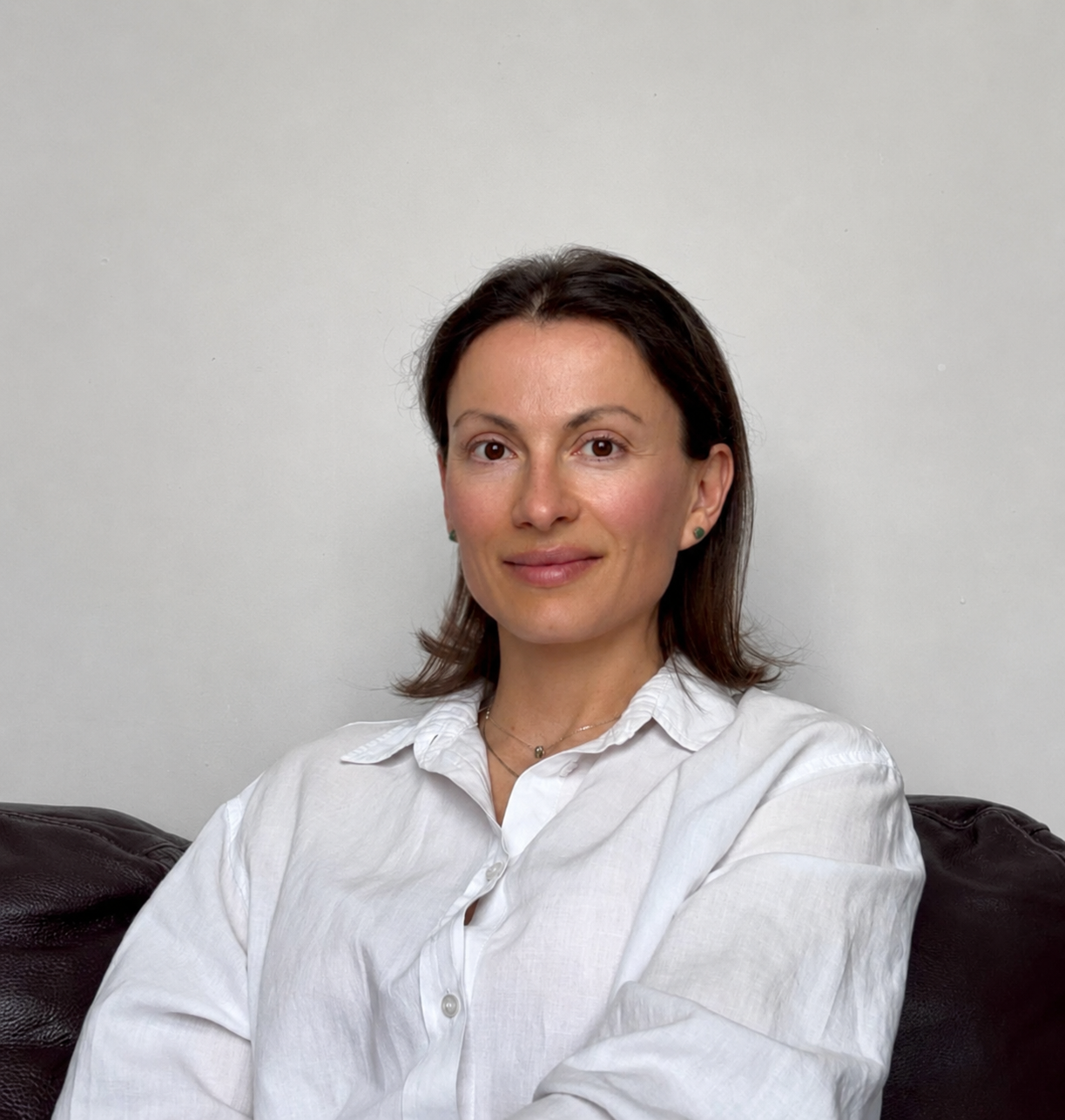 A woman with dark brown hair, wearing a white button-up shirt, sitting on a dark-colored couch against a plain white wall.