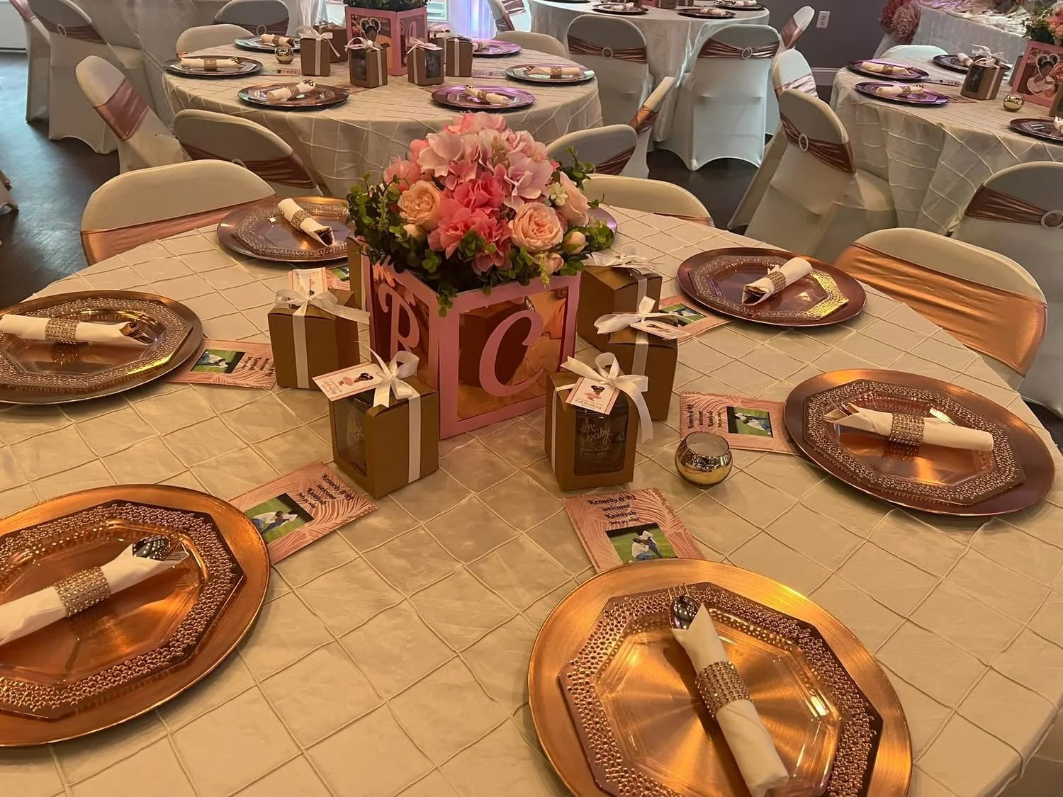 A banquet table decorated with a pink floral centerpiece, gold and pink wrapped gifts with white ribbons, pink and clear plates, gold napkins, and pink placemats, set for a celebration.