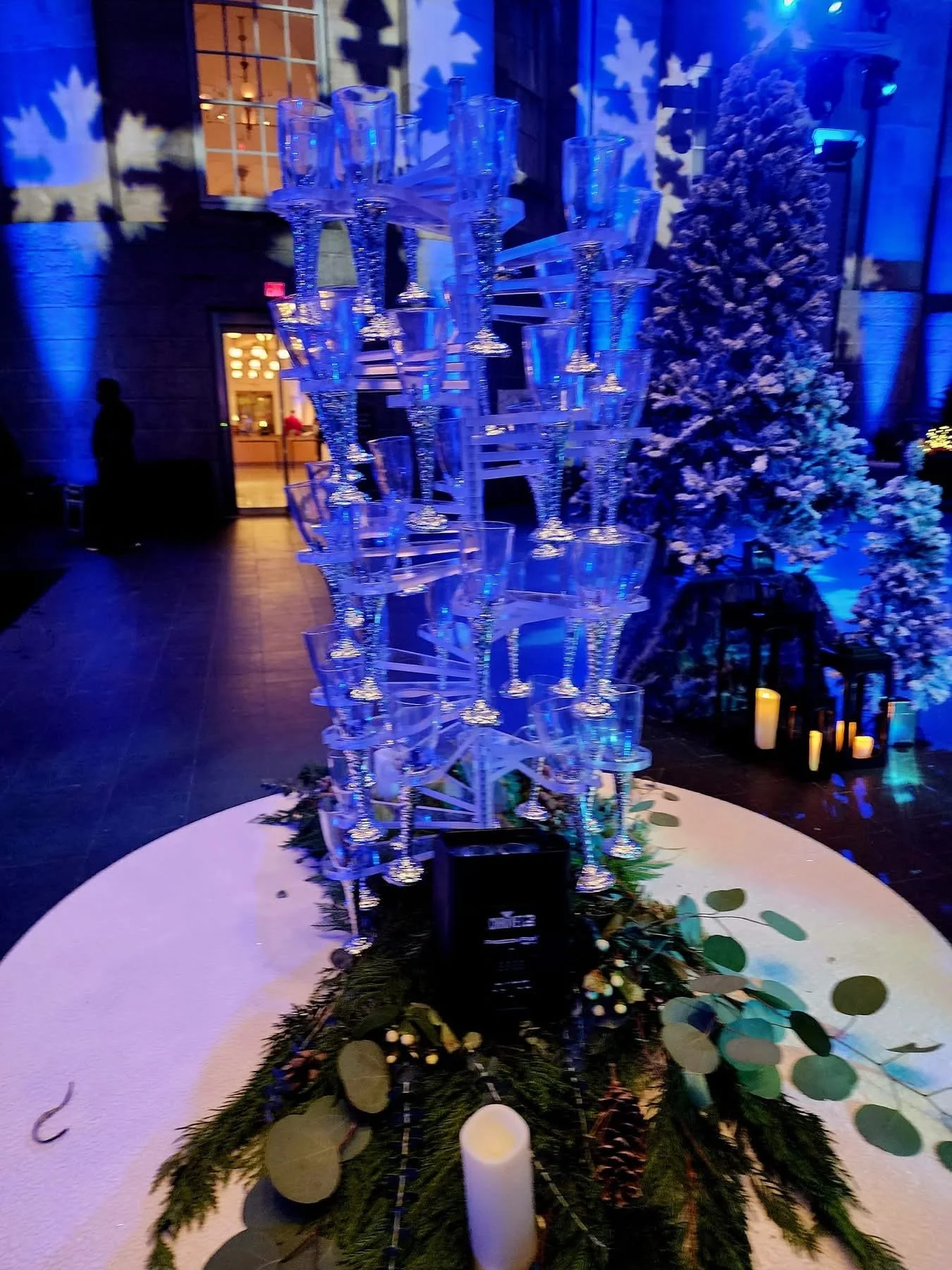 A decorative display of numerous clear glass champagne flutes arranged in a tall, artistic sculpture on a table decorated with greenery and candles, with a winter-themed background including a snow-covered artificial Christmas tree and ambient blue l