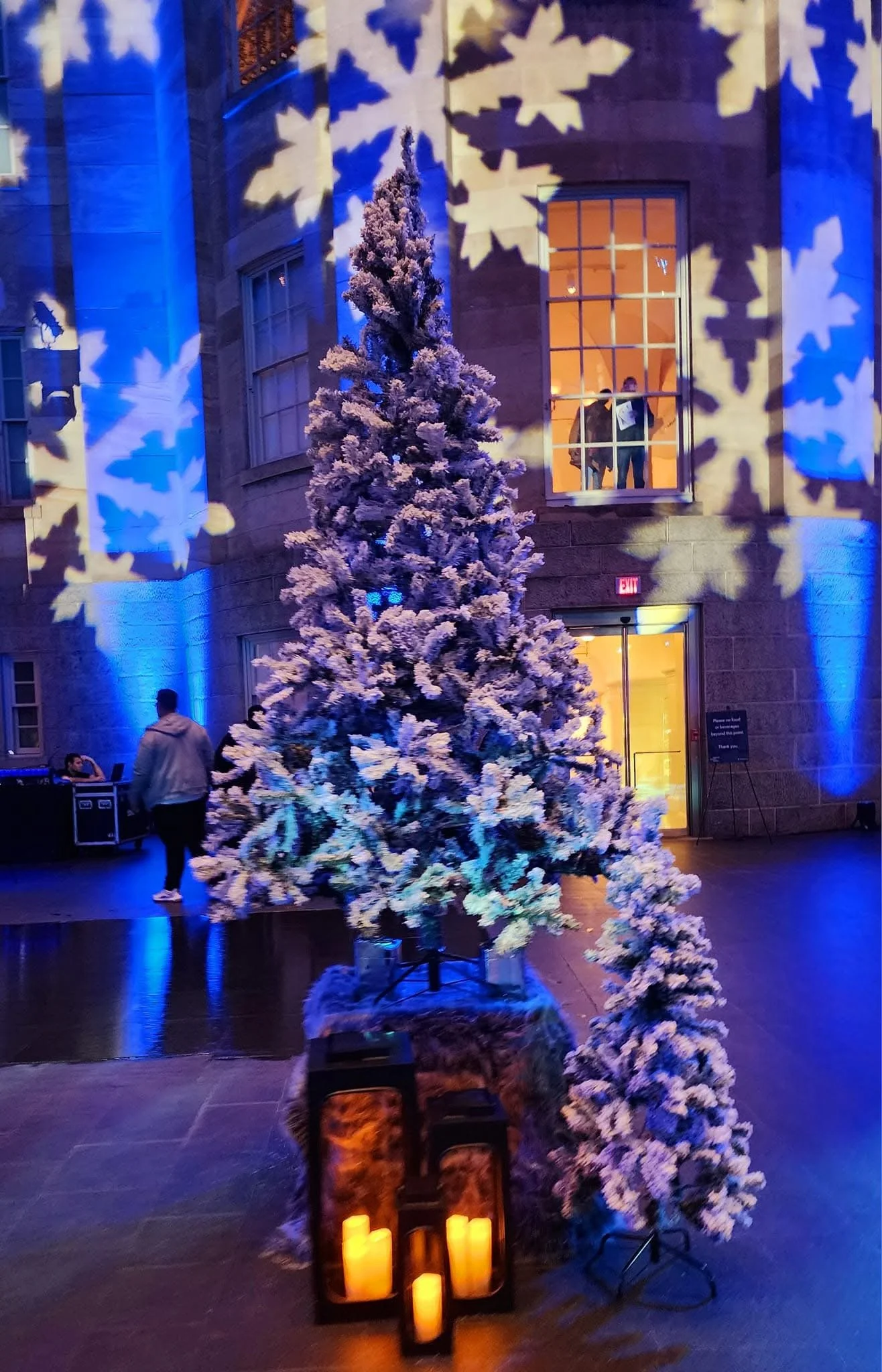 A decorated Christmas tree with artificial snow, lit candles in lanterns at the base, and blue and white snowflake projections on the wall behind, inside a building with warm lighting.