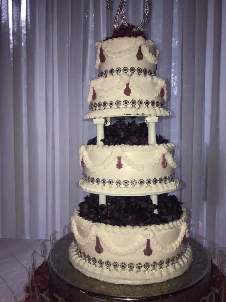 A large, multi-tiered wedding cake with white icing, black decorative accents, and purple embellishments, topped with a silver decorative topper, displayed in front of a light-colored curtain backdrop.