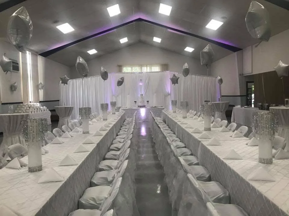 A banquet hall decorated in white for a celebration, featuring long tables with white tablecloths, white napkins, and chairs covered with white cloth. Silver balloons are floating above, and tall centerpieces with crystal embellishments are arranged 
