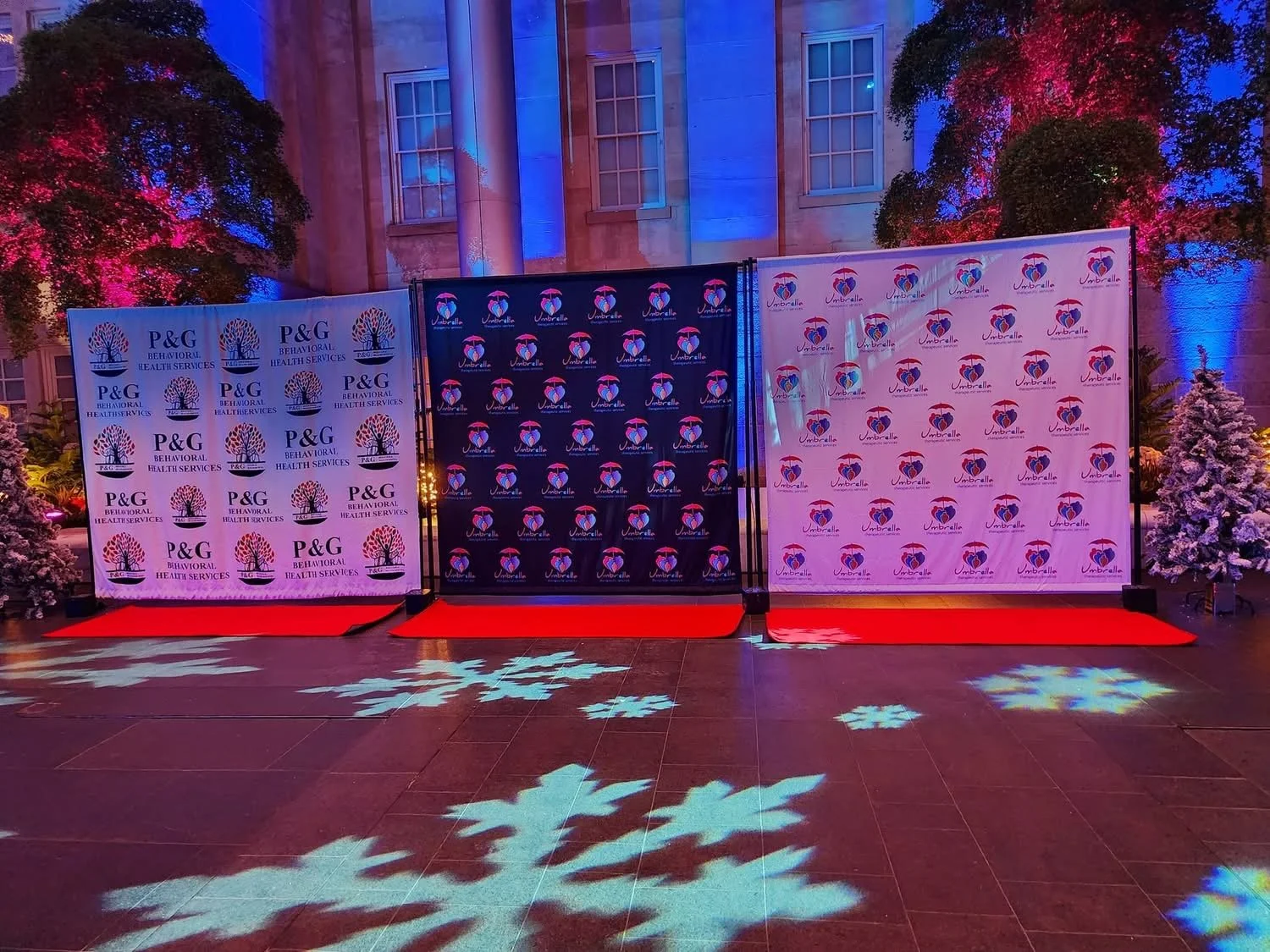 Three backdrop banners with logos for P&G Behavioral Health Services, Umbrella, and another unidentified logo, set on red carpet, with colorful lighting illuminating trees and wall in the background.