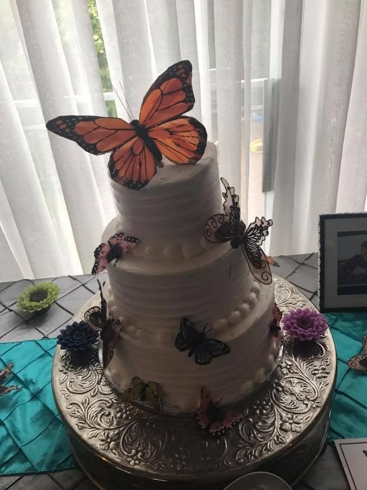 A three-tiered white wedding cake decorated with colorful butterfly toppers and embellishments, placed on a decorative silver tray.
