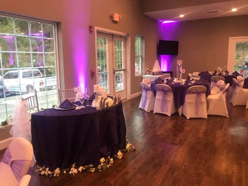 Decorated banquet hall with round tables covered in purple tablecloths, white chairs with purple sashes, a wedding cake, and a table with floral arrangements, illuminated with purple lighting.