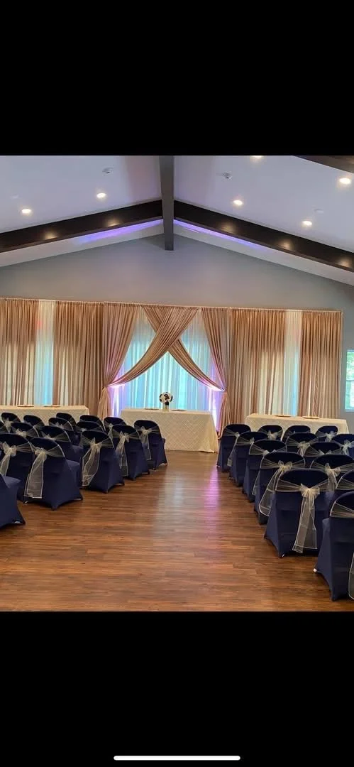 Event hall setup with rows of chairs covered in navy blue fabric with gold bows, a decorated table at the front with beige curtains and soft lighting, wooden floor and a peaked ceiling with recessed lights.