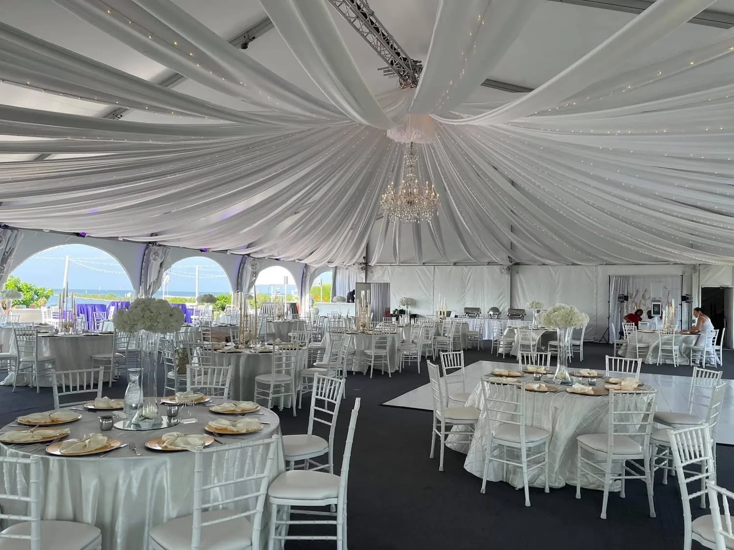 Elegant event tent decorated with white drapery and crystal chandeliers, featuring multiple round tables set with white tablecloths, floral centerpieces, and place settings, overlooking a view of the ocean in the background.