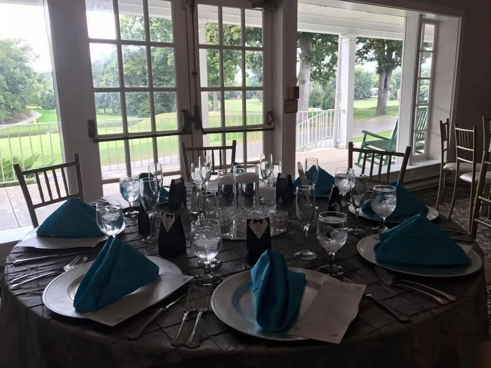 A round dining table set for a formal event with blue napkins, wine glasses, water glasses, and black and white decorations, situated indoors near large windows with a view of a golf course outside.