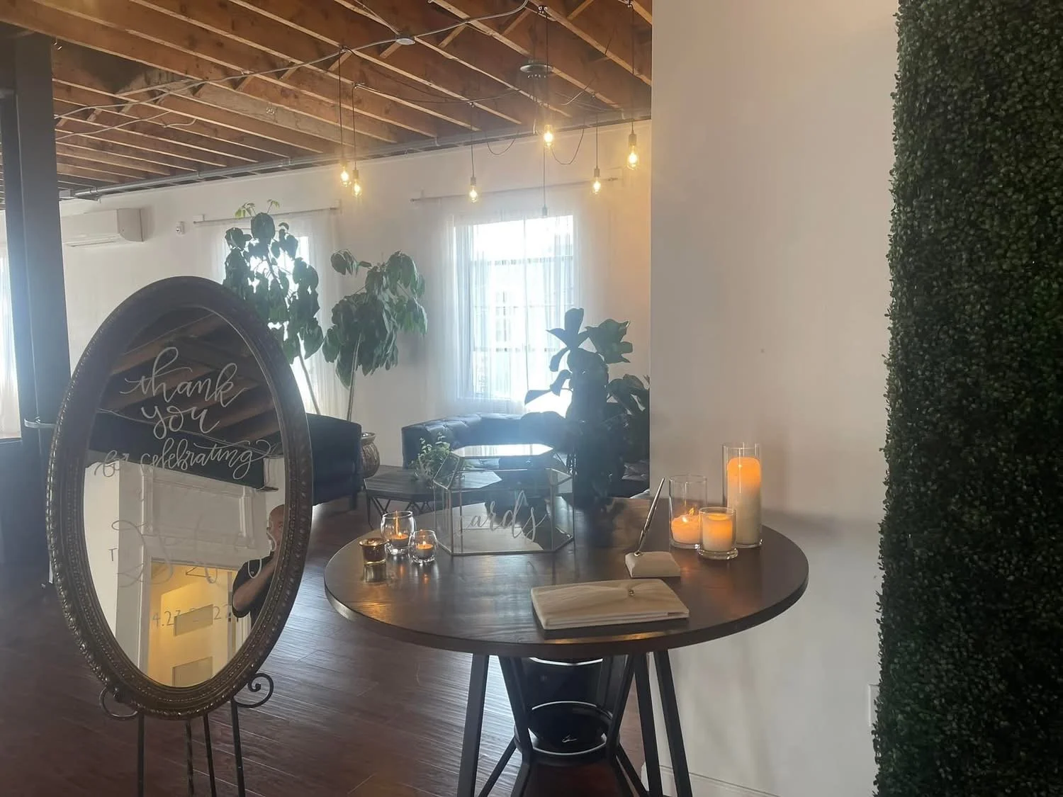 Interior of a decorated event space with a large oval mirror with 'thank you for celebrating' written on it, candles on a round table, potted plants, and seating area with a window in the background.
