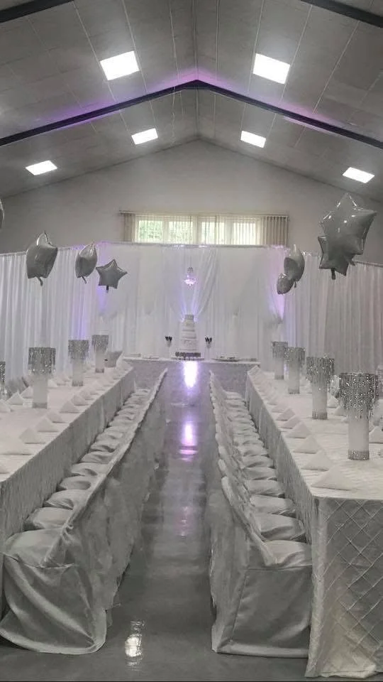 A white-themed banquet hall decorated with star-shaped balloons, elegant table settings, and a cake at the front, likely set up for a celebration or wedding reception.