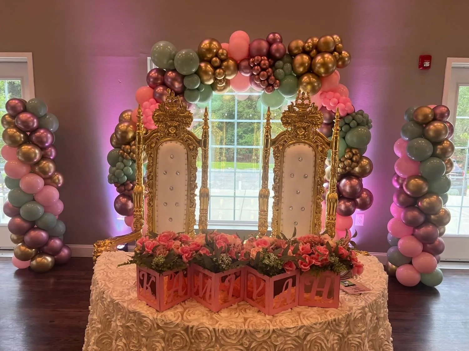 Royal celebration setup with two ornate gold and white thrones, a floral arrangements, and a balloon arch with pink, green, and purple balloons in a decorated room.