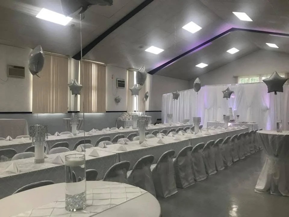 Decorated banquet hall with long tables covered in white tablecloths, star-shaped balloons, floral centerpieces, and a draped backdrop with purple lighting for a celebration or event.