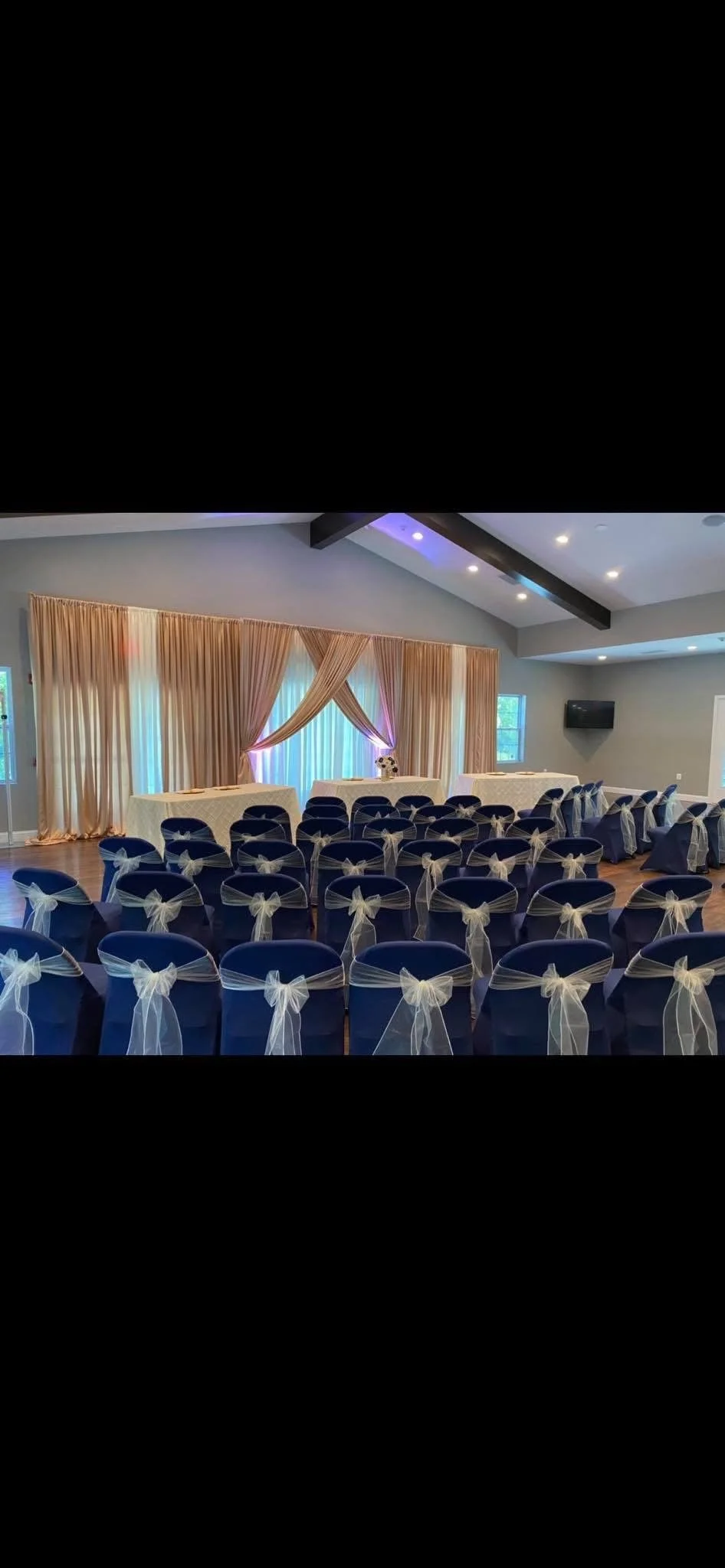 Wedding ceremony setup with rows of blue chairs adorned with white bows facing a decorated table and curtains.