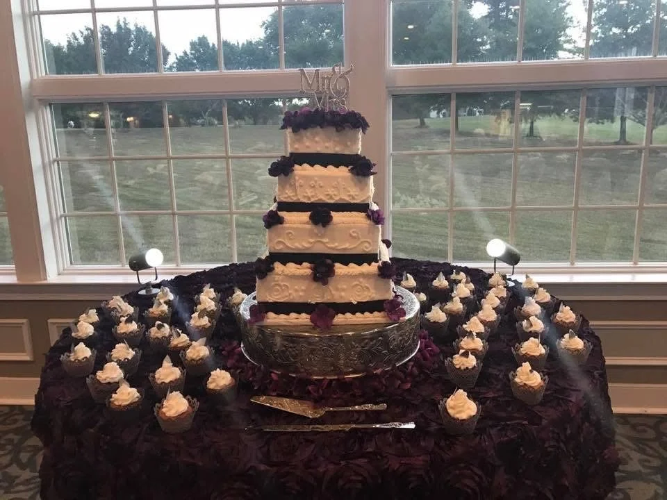 A wedding cake display with a tall, multi-tiered cake decorated with purple and black flowers, surrounded by cupcakes with white frosting on a table covered with a dark cloth, in front of large windows overlooking a grassy yard.