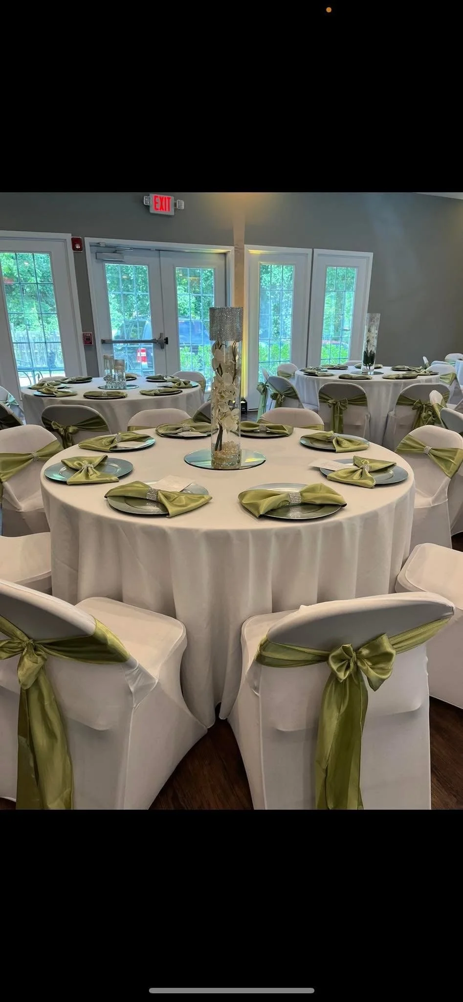 Elegant banquet hall with round tables covered in white tablecloths, each set with chairs tied with green sashes, and decorated with tall glass centerpieces, in a room with large windows and glass double doors.