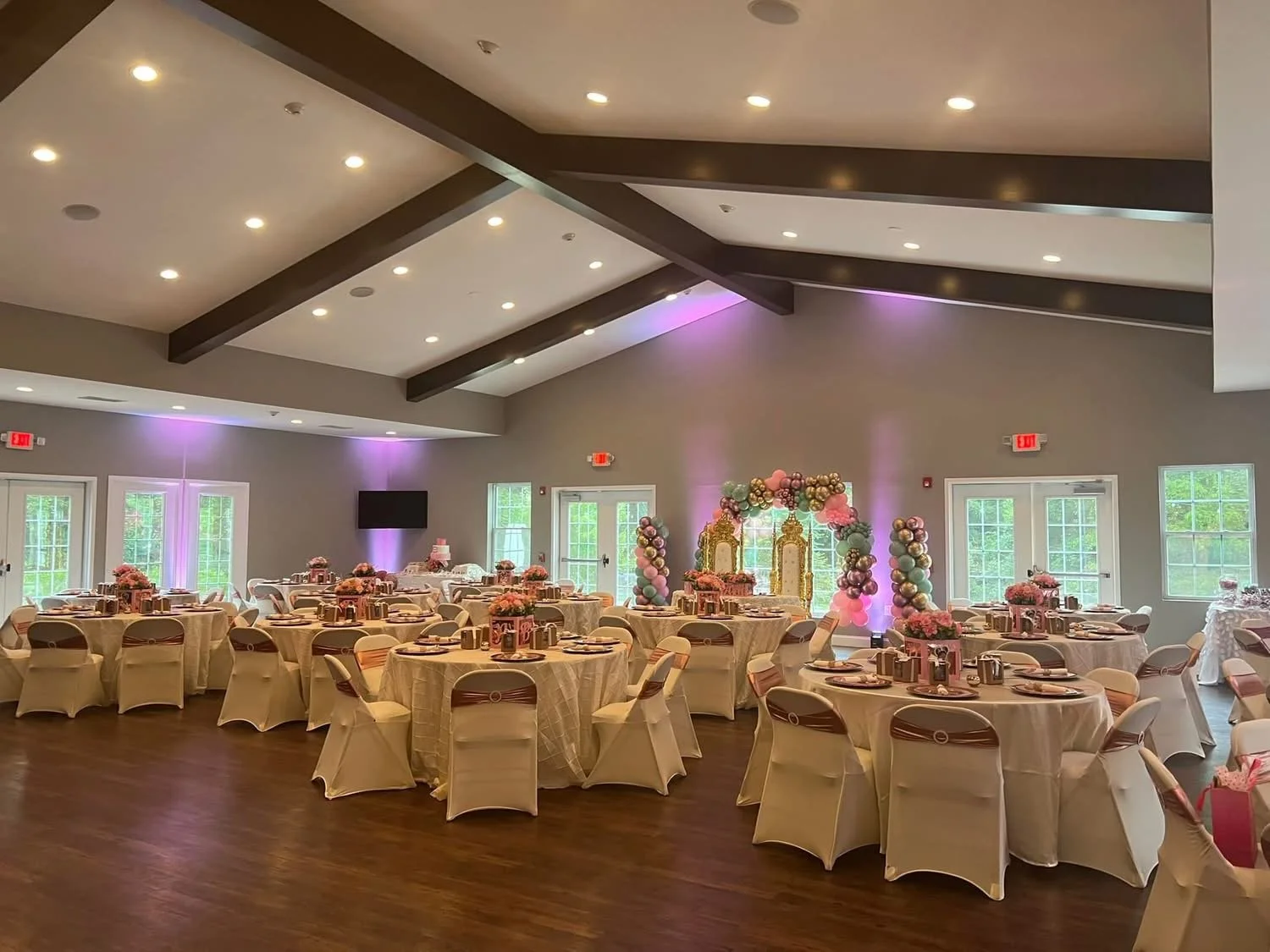 A decorated banquet hall with round tables covered in white tablecloths, arranged with pink floral centerpieces, set with plates and silverware, and surrounded by white chairs with ribbons. The background features a balloon arch and pastel-colored de