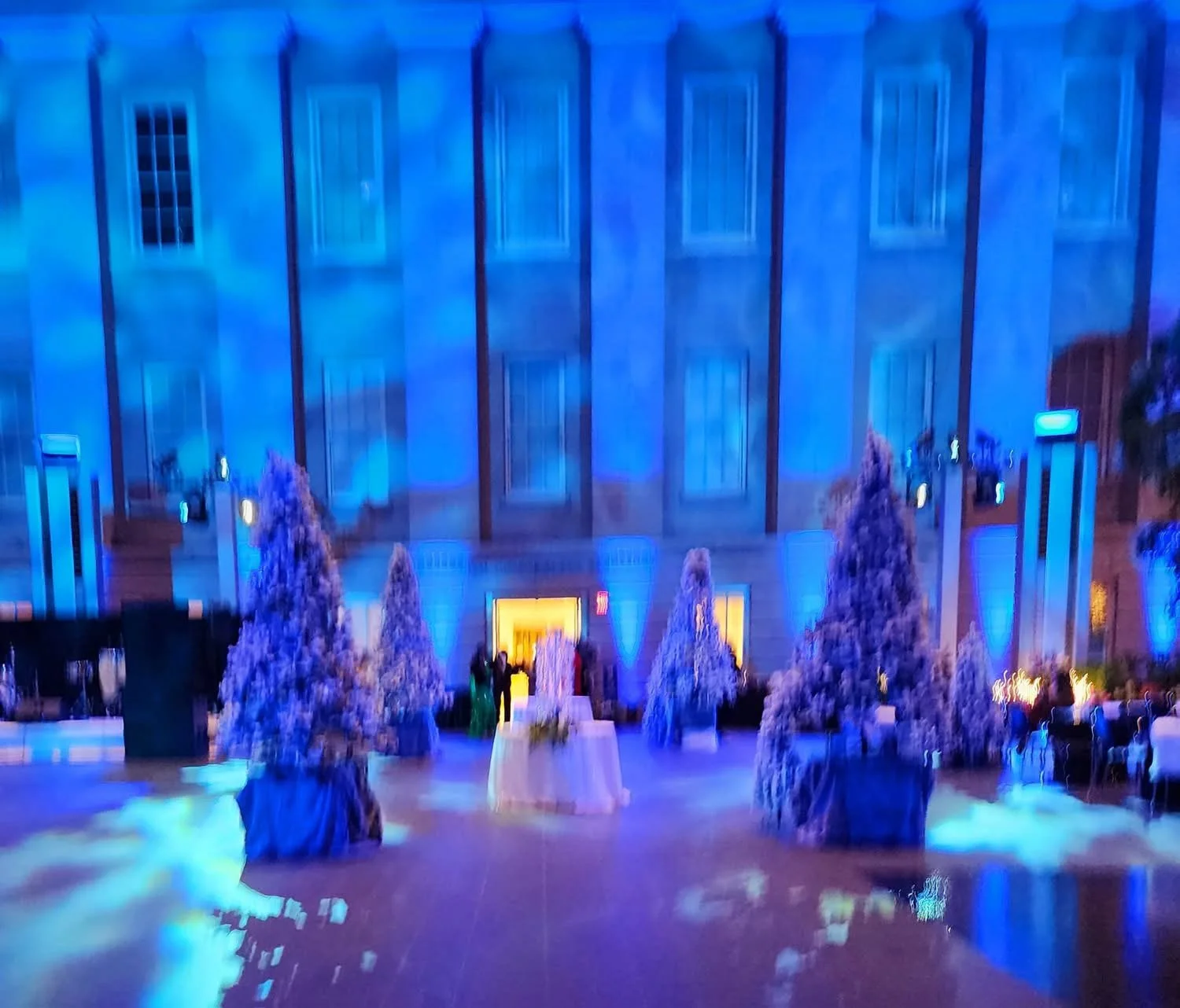 The image shows a dimly lit indoor space decorated with blue lighting, purple frosted pine trees, and some people gathered near a table in front of a building with multiple windows and vertical pillars.
