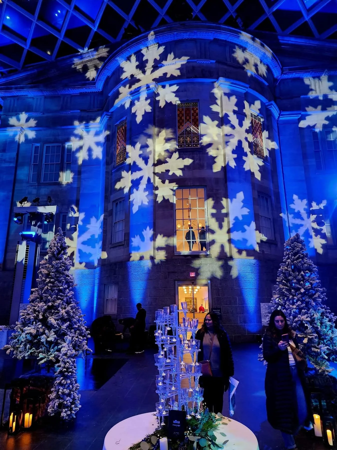 Indoor scene with Christmas and winter holiday decor. Large snowflake projections on the building's facade, two snow-covered trees, and people walking around, including a woman looking at her phone. There is a table with a tall glass candle holder ce