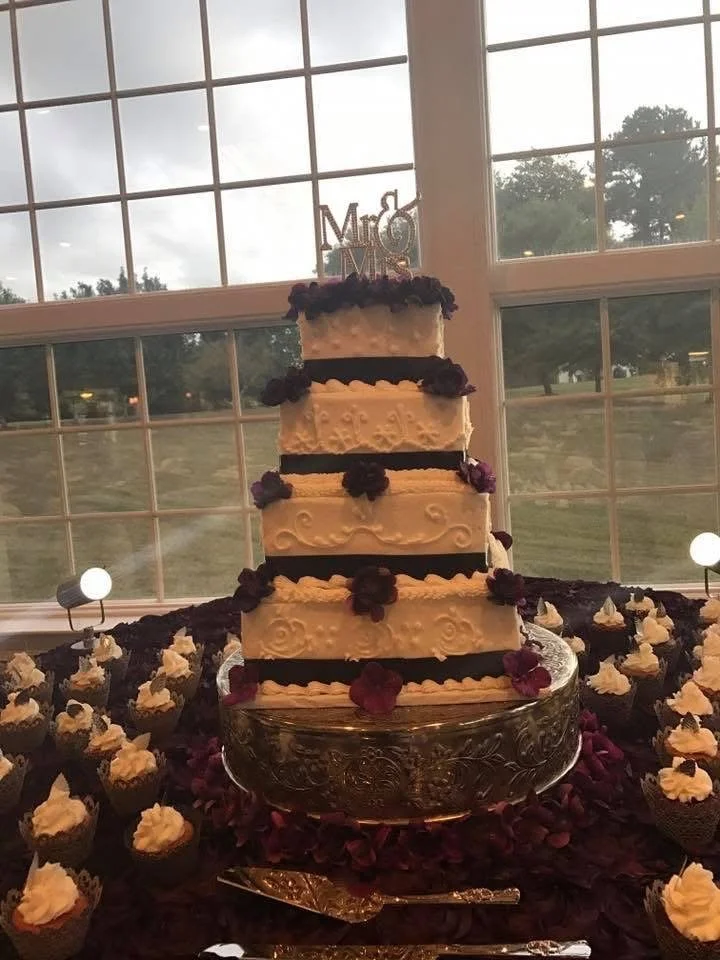 A four-tier wedding cake decorated with purple flowers and a 'Mrs & Mr' topper, surrounded by cupcakes with white frosting, set on a table with a black cloth and rose petals, in front of a large window showing an outdoor scene.