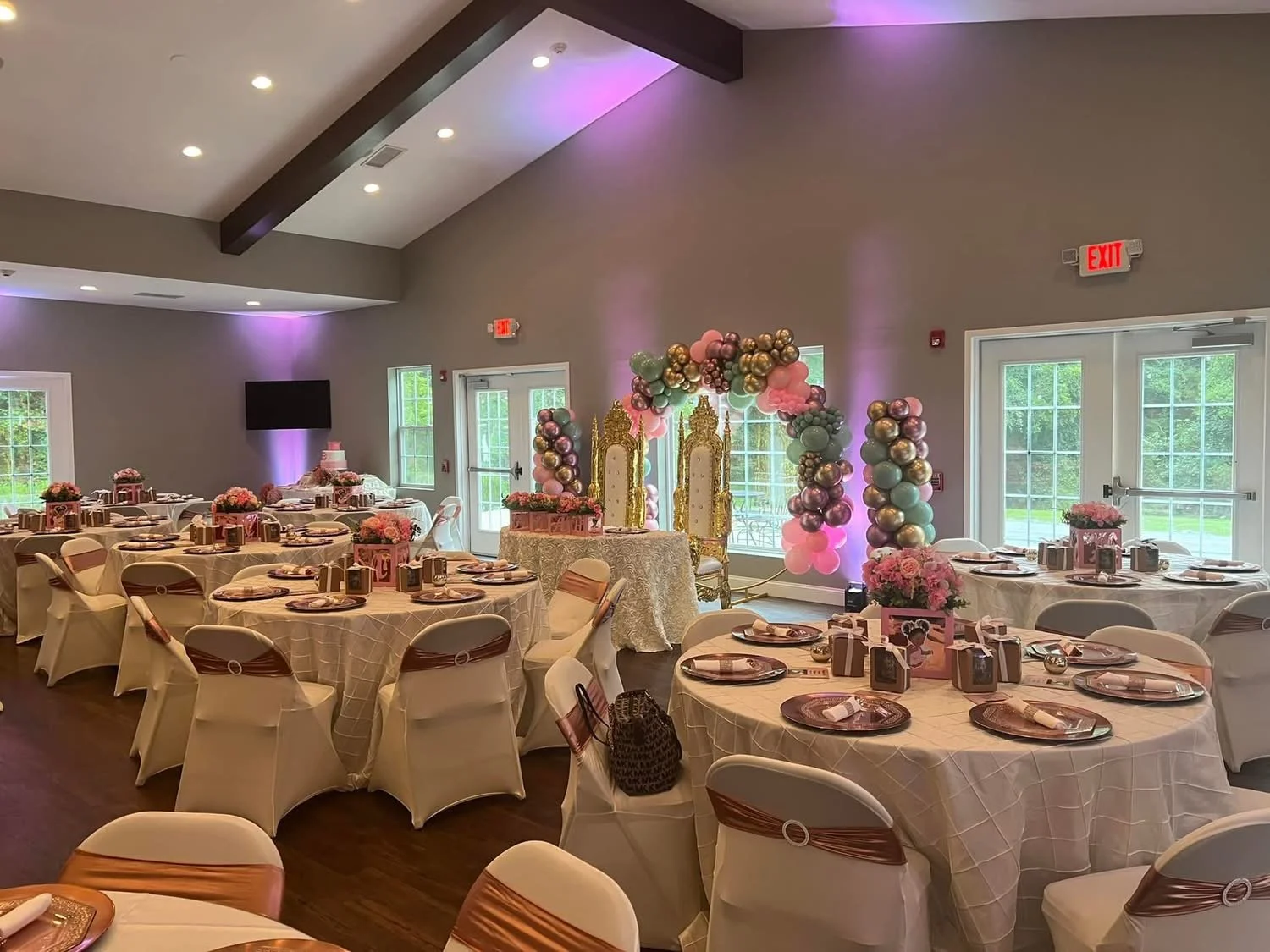 Decorated event space with round tables covered in pink and white tablecloths, pink accents, and balloon arch with pink, gold, and green balloons, featuring two ornate golden chairs underneath.