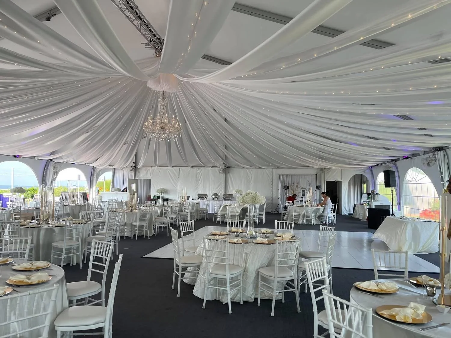 Elegant event tent interior with white draped ceiling, chandelier, round tables with white tablecloths, floral centerpieces, and white chairs, set for a formal occasion.