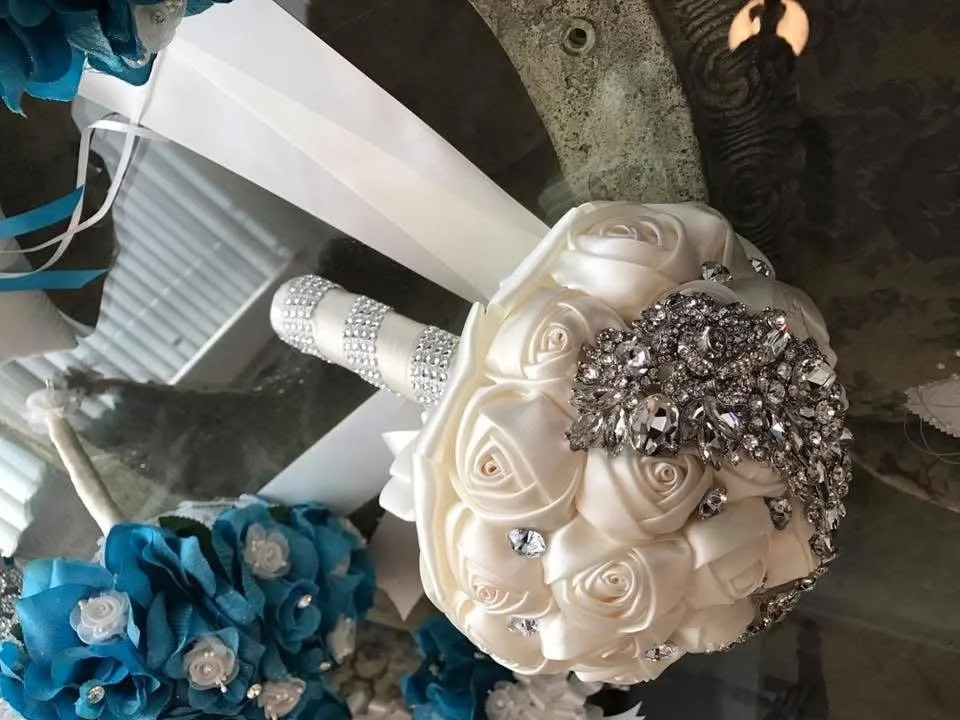 A decorative bouquet of white roses with a band of rhinestones, placed on a surface near a mirror, with a pair of blue fabric flowers.