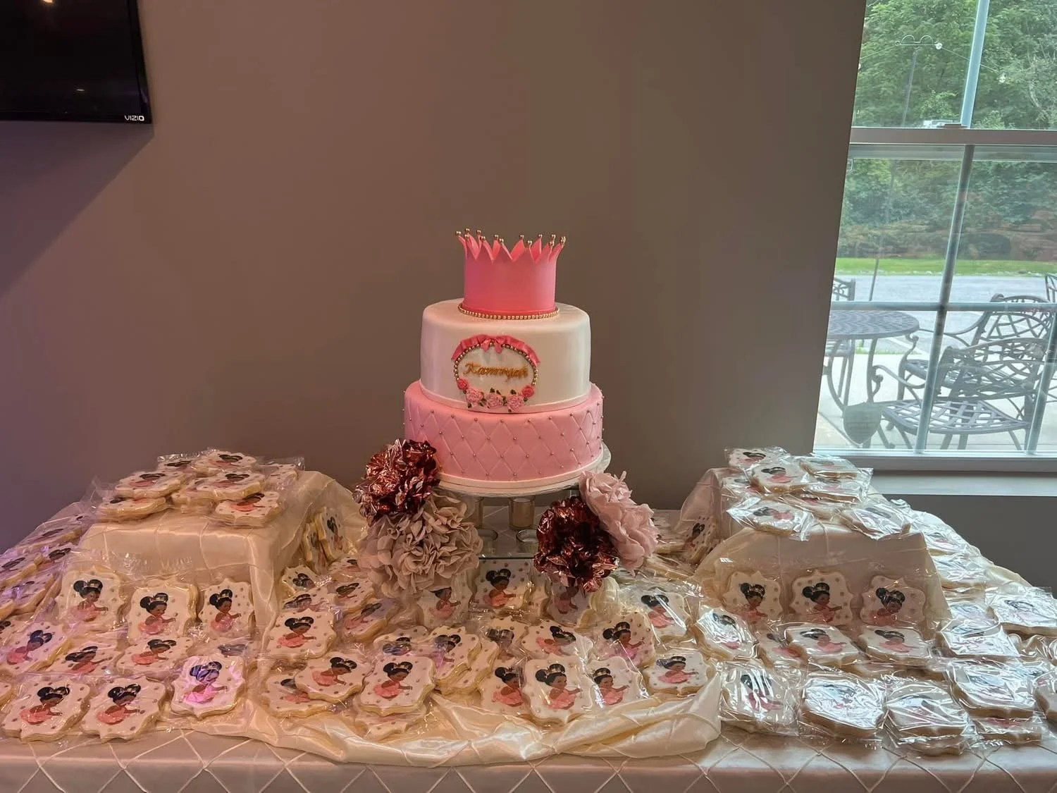 A three-tier pink and white birthday cake with a pink crown topper and floral decorations, surrounded by decorated cookies and flowers on a table.