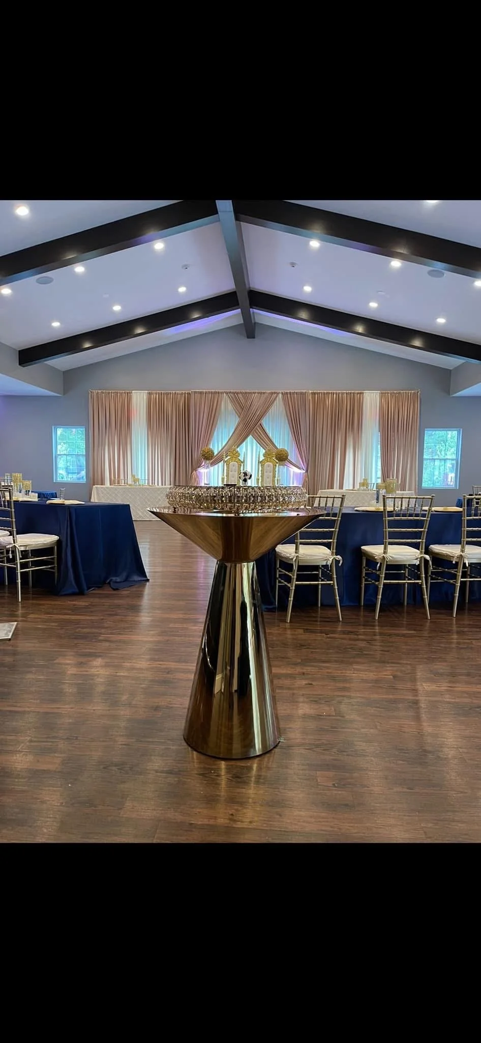 Elegant event space with a high ceiling, decorated with draped peach-colored curtains and floral arrangements, featuring a table with champagne glasses in the foreground and tables with navy blue tablecloths and gold chairs in the background.