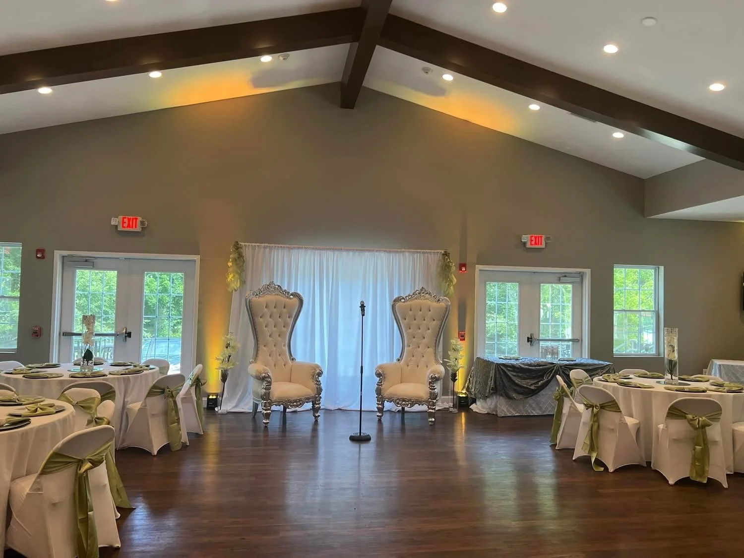 Event space decorated with two large white high-back chairs, a backdrop with white drapery, and round tables with white tablecloths and gold sashes on chairs. The room has wooden floors, large windows, and ceiling lights.