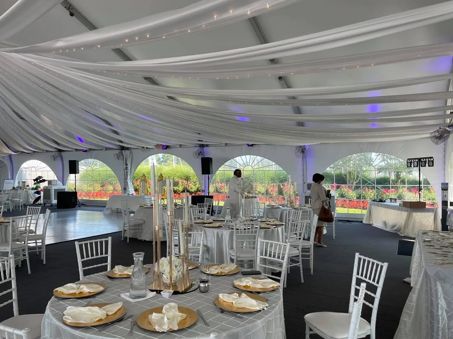 Elegant event tent with white draped ceiling, decorated round tables with white chairs, and large windows showing outdoor garden with vibrant flowers.