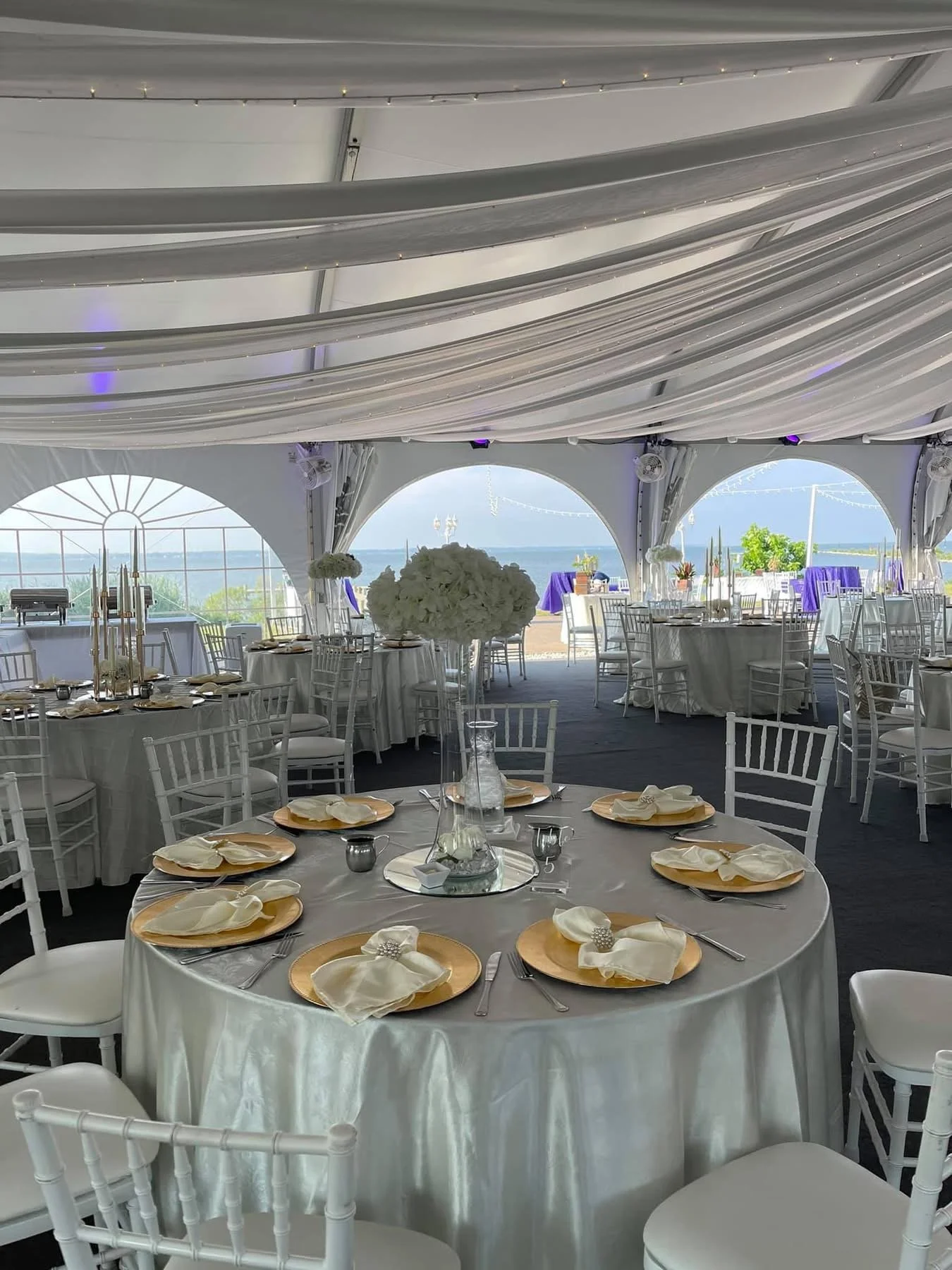 Indoor event space decorated for a wedding or celebration with round tables, white chairs, elegant table settings, and a view of the water outside.