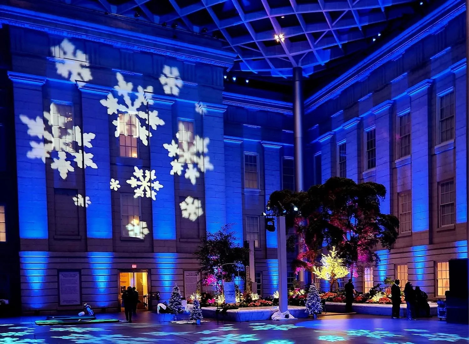 Indoor scene with blue lighting, snowflake projections on the walls, and Christmas trees decorated with lights and ornaments. Several people are standing and walking around in the space.