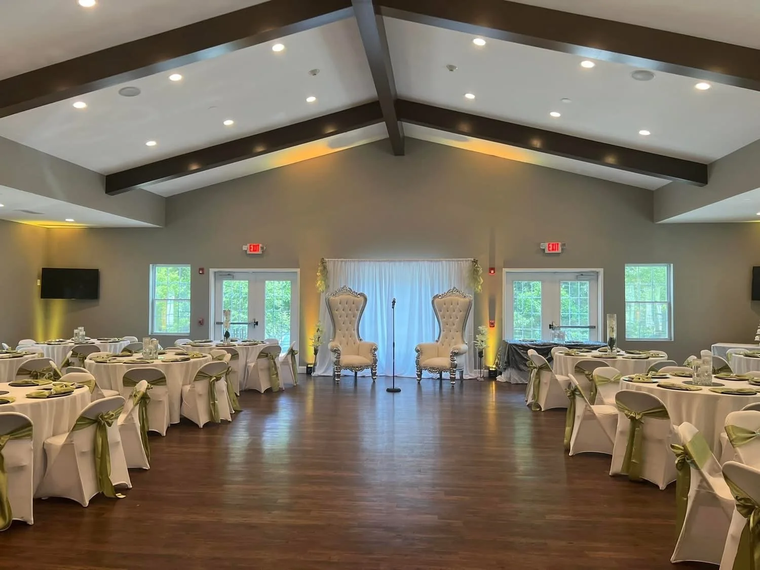 Wedding reception hall with round tables covered in white tablecloths, decorated chairs with green sashes, and a small stage area with two ornate high-backed chairs and a microphone. There are windows, a door, and TVs on the walls.