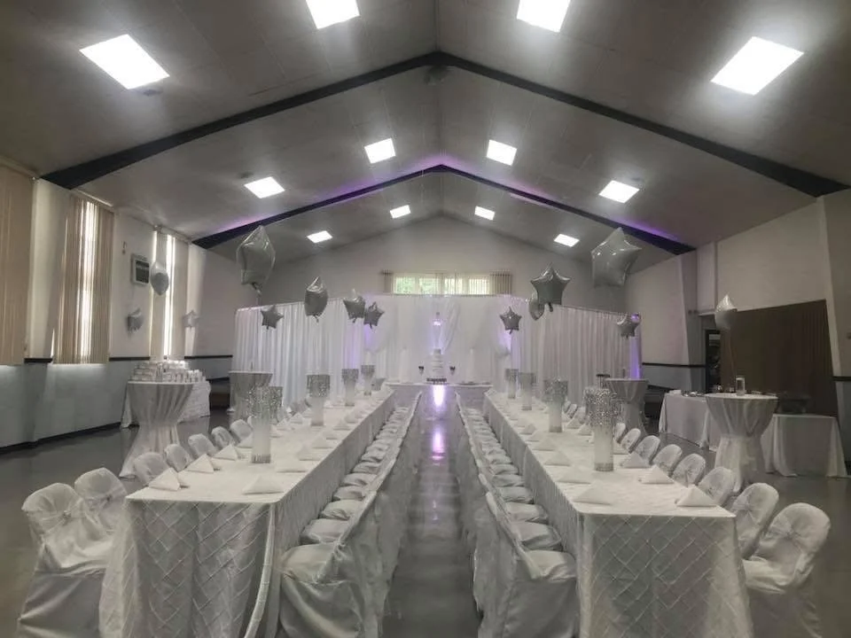Elegant banquet hall decorated for a wedding reception, with long tables draped in white, star-shaped balloons, and a backdrop with white curtains and purple lighting.
