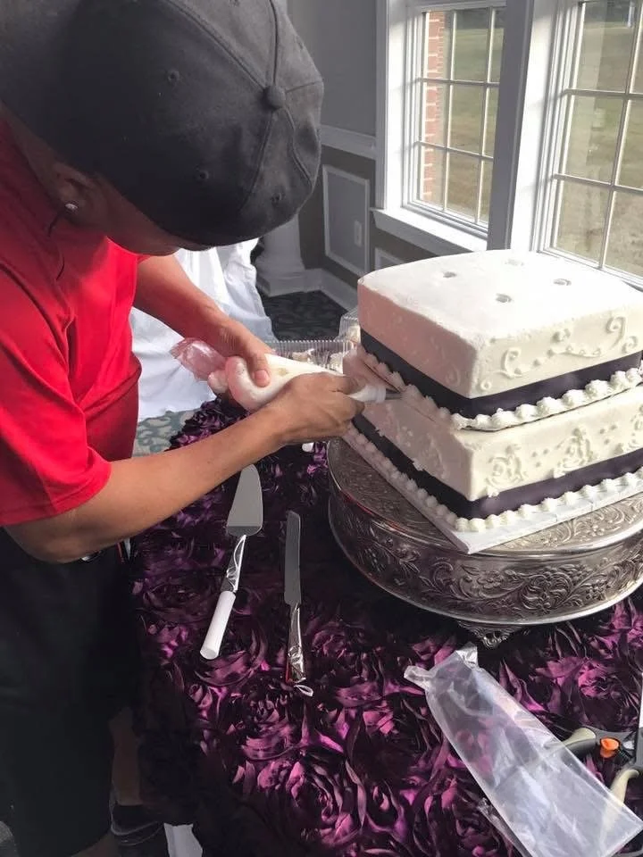 Person wearing a gray cap and red shirt decorating a large, tiered wedding cake with white icing and black accents, placed on an ornate silver stand near a window.