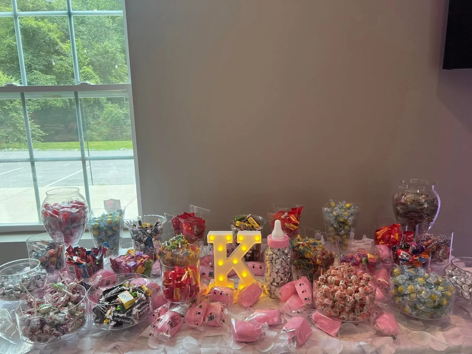 Candy table with jars of various candies, pink decorations, and a lit letter 'K' centerpiece, set near a window with green trees outside.