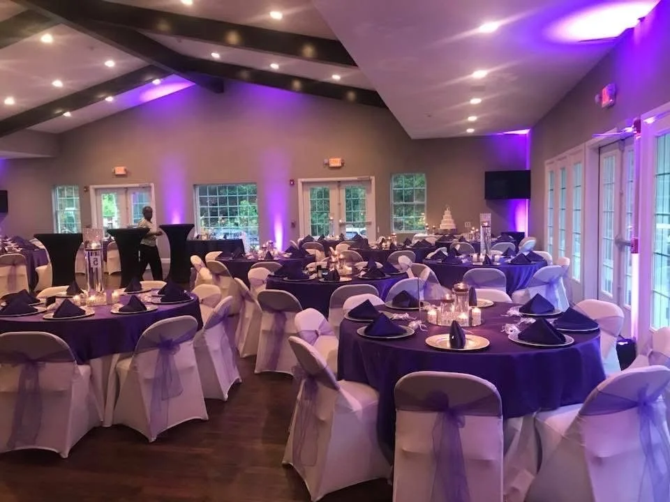 Elegant event room decorated with purple and white tables and chairs, black napkins, purple lighting, and a scenic view outside through windows.