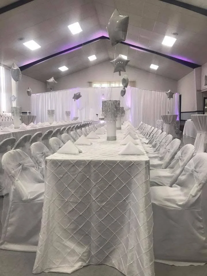 Elegant banquet hall decorated in white, with long tables covered in quilted white tablecloths, white chairs with bows, and star-shaped balloons hanging from the ceiling.