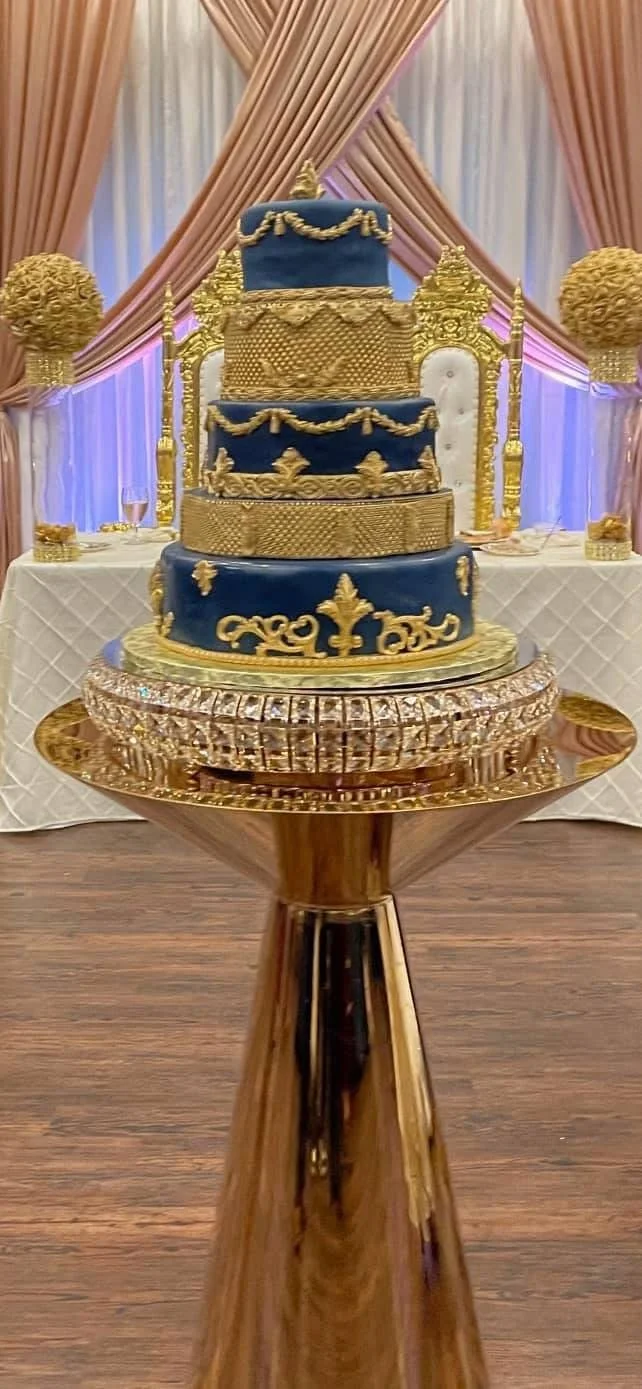 A multi-tiered wedding cake with blue and gold decorations, set on a gold and crystal cake stand on a wooden table in a decorated event space.