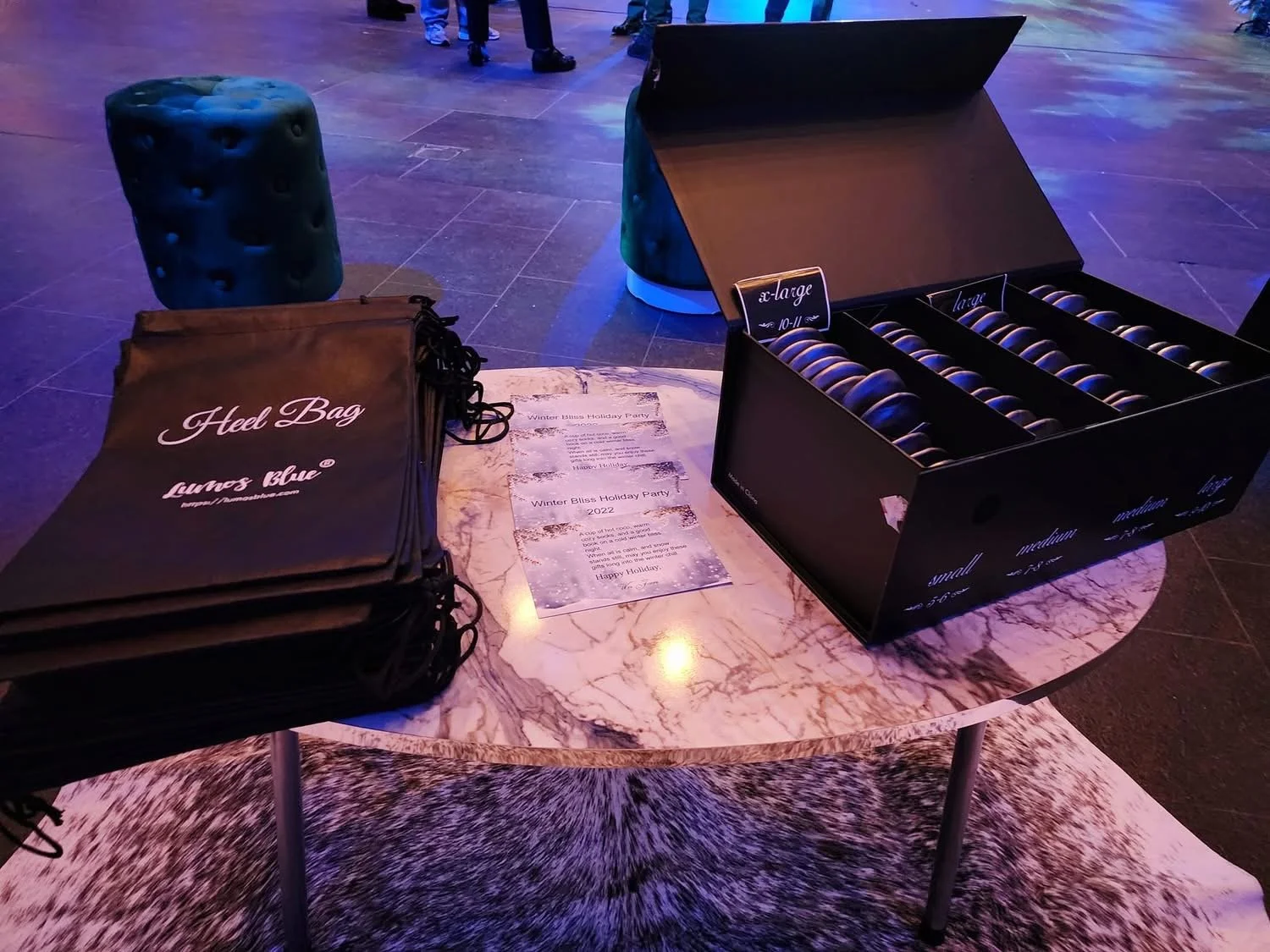 Table with black shoe bags, a box of black shoes, and printed event flyers, in a dimly lit venue with purple and blue lighting.