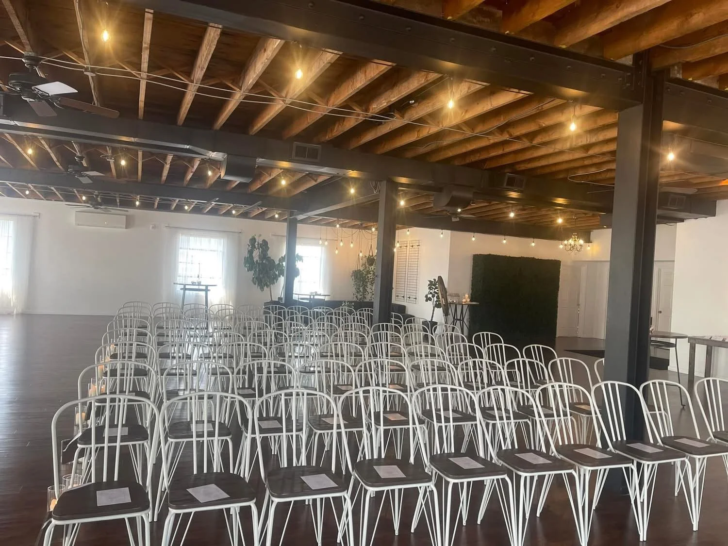 An event space with rows of empty white chairs with black seats arranged facing a small stage or focal point. The room features exposed wooden beams on the ceiling, string lights, and a few potted plants near windows.