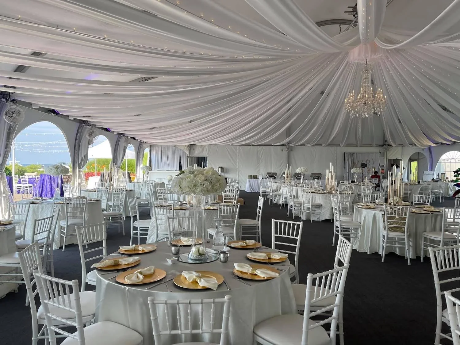 Elegant event tent with white drapery ceiling, chandelier, round tables with white tablecloths, floral centerpieces, and gold-rimmed plates, set for a formal celebration.