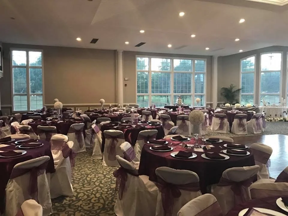 Event hall decorated with round tables covered in burgundy tablecloths, white chairs with burgundy sashes, white floral centerpieces, and table settings with plates, napkins, and glasses, ready for a formal celebration.