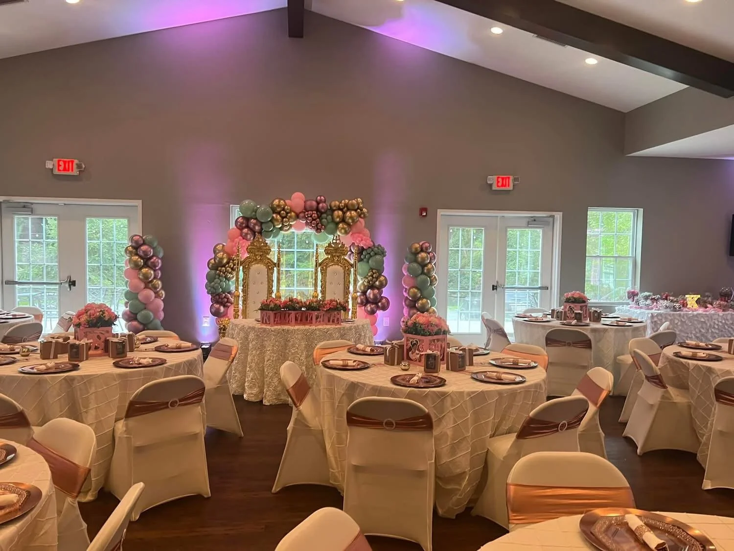 Decorated event hall with round tables covered in beige tablecloths and white chairs with brown sashes. A pink, purple, gold balloon arch and gold throne chairs on a table serve as the focal point, indicating a celebration or party.