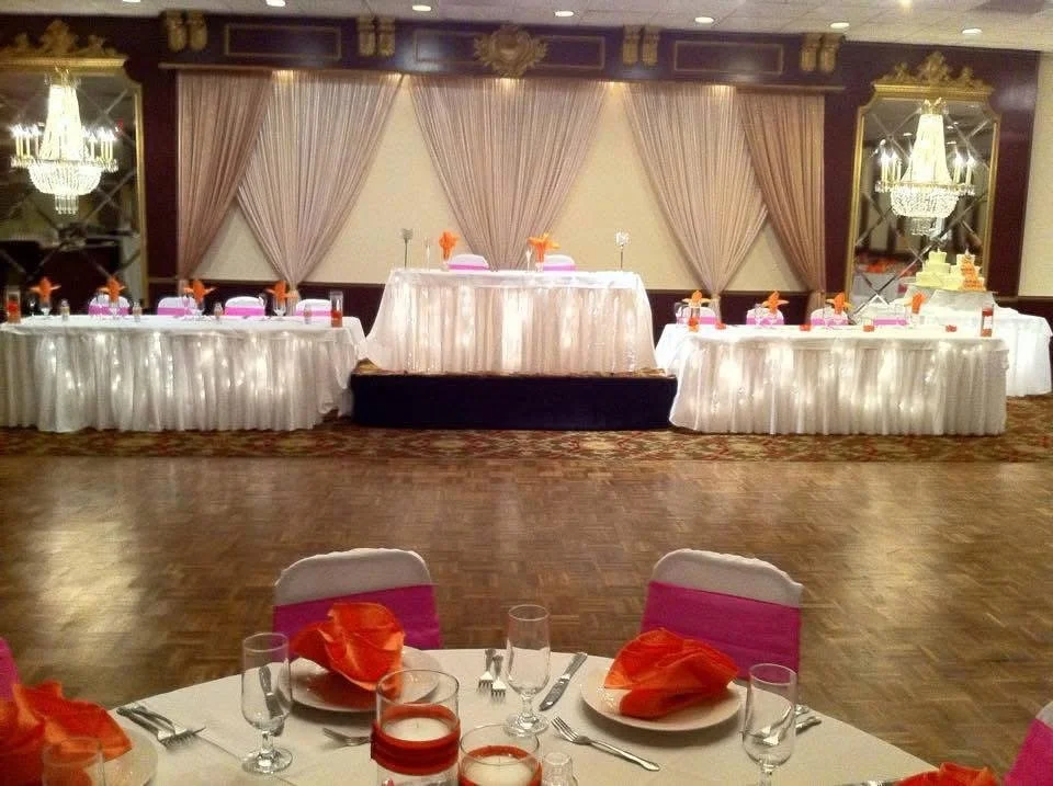 Banquet hall decorated for a wedding reception with a head table, wedding cake, and table settings with pink and orange accents.