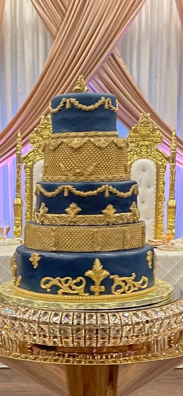 A multi-tiered cake decorated with navy blue and gold, featuring ornate gold designs and garlands, placed on a gold stand in front of beige and pink drapery.