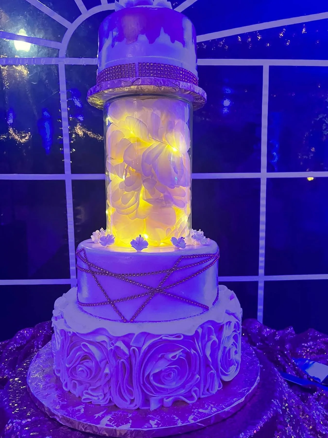 Three-tiered birthday cake with purple and white decorations, illuminated with purple and yellow lighting, decorated with roses made of frosting on the bottom layer, chains of beads on the middle layer, and a glass display with a hat-shaped top on th