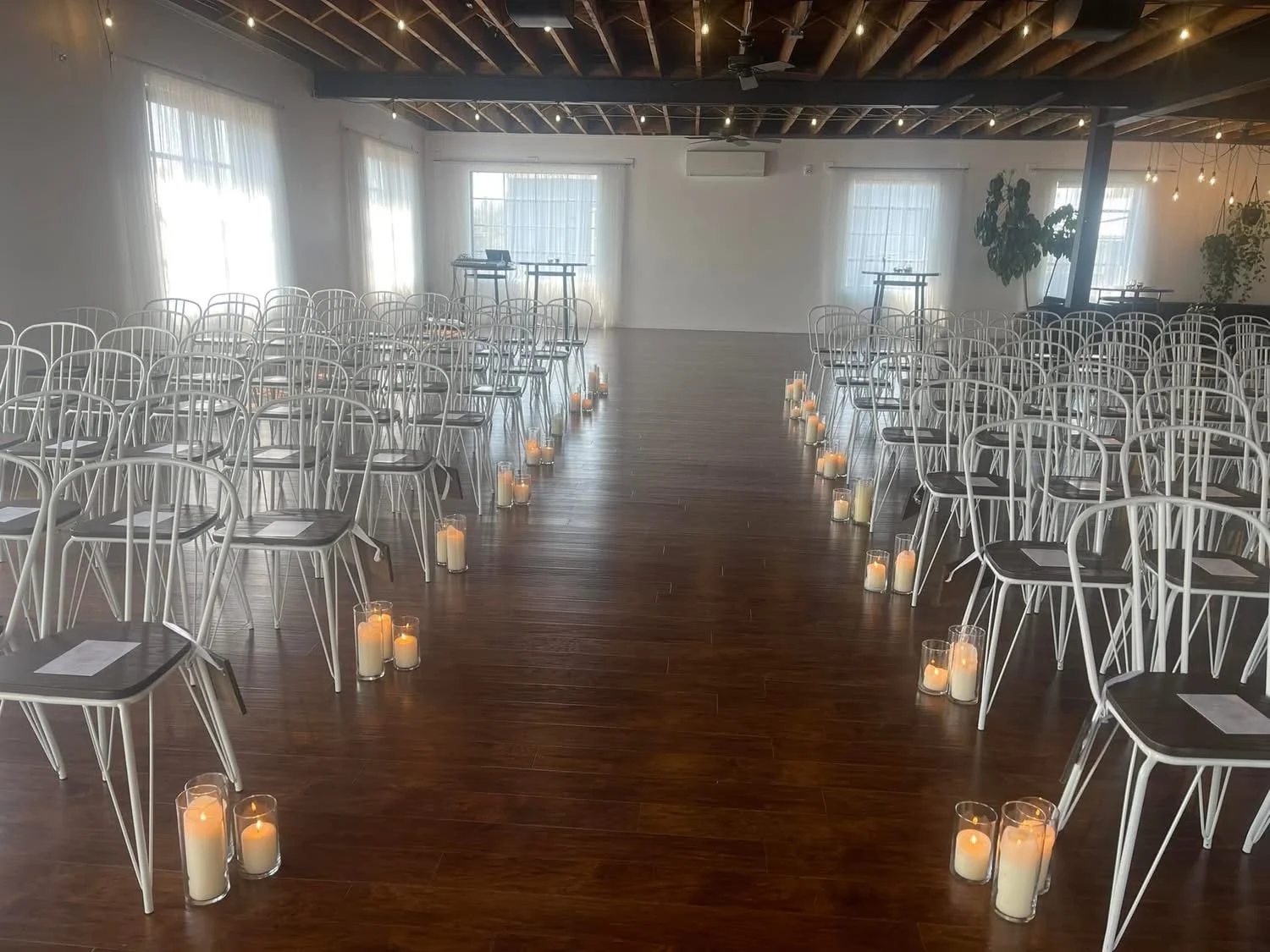 An indoor event space with rows of white chairs arranged along a wooden aisle, which is lined with lit candles in glass holders. The room has white walls, large windows with sheer curtains, and ceiling beams, creating an elegant setting for a ceremon